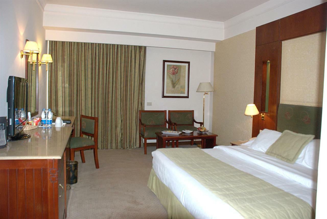 Safir Hotel Cairo Giza Quarto foto The photo shows a hotel room. It features a bed with white linens and a green throw. On one side of the bed, there's a small bedside lamp. Opposite the bed is a desk with a mirror above it, alongside a couple of chairs and a small table. The room has