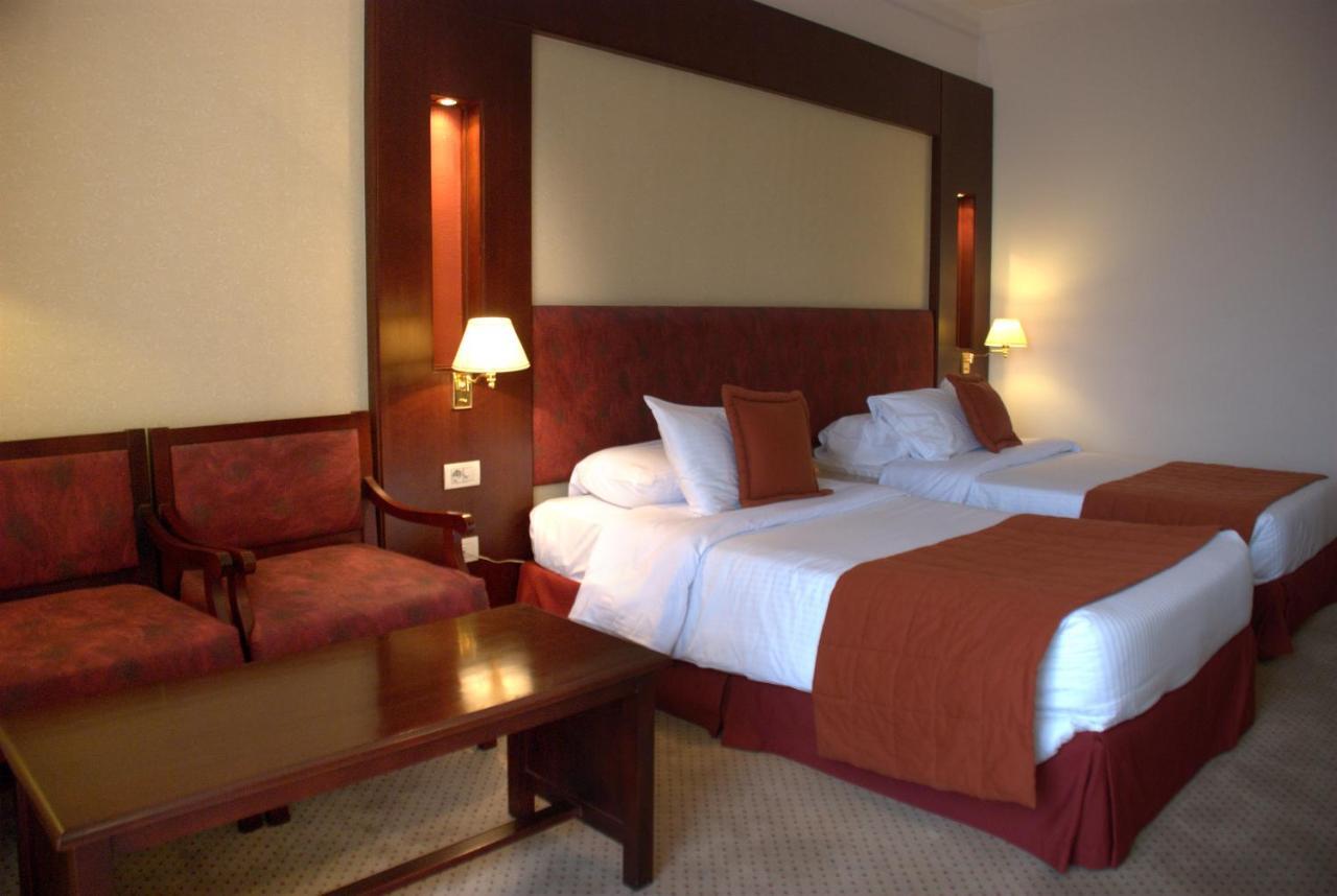 Safir Hotel Cairo Giza Exterior foto The photo shows a hotel room featuring two twin beds with white linens and orange bedspreads. There is a headboard behind the beds, along with wall-mounted lamps on either side. In the corner, there are two upholstered chairs and a wooden coffee tabl
